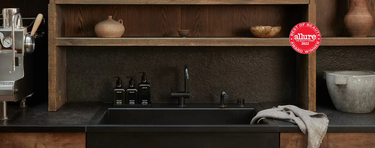 Three black pump bottles on a tray beside a matte black kitchen sink under wooden shelves with an Allure Best of Beauty 2022 award badge visible